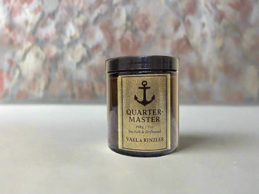 Candle of 'Quarter Master' by Vael & Rinzler on a white surface with a blurred floral background-luxury religious scented candles with label and gold background