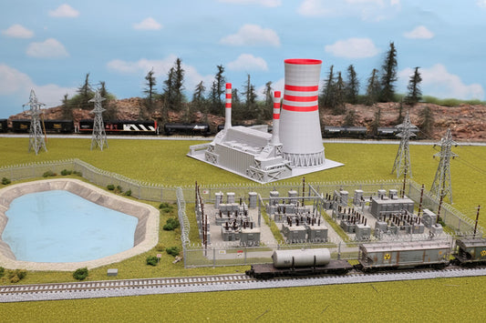 A Z scale model of a power plant featuring a large cooling tower with red and white stripes, placed on a green surface, possibly representing grass, with a model train and trees in the background.