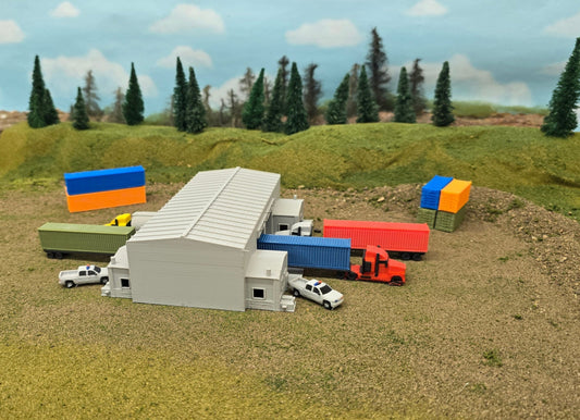 Model of a warehouse with colorful shipping containers and trucks on a grassy field with trees in the background.