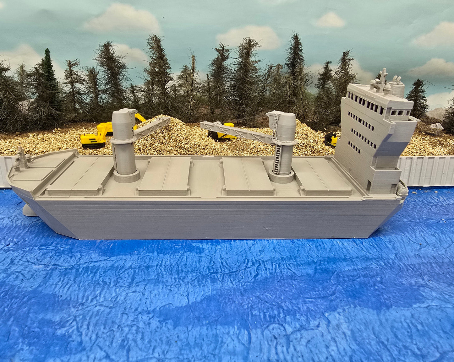 Model of a large ship on a blue surface with trees and construction vehicles in the background