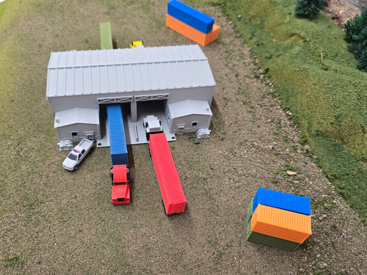 Model of a train station with a gray building, red and blue train cars, and a white car on a grassy field.