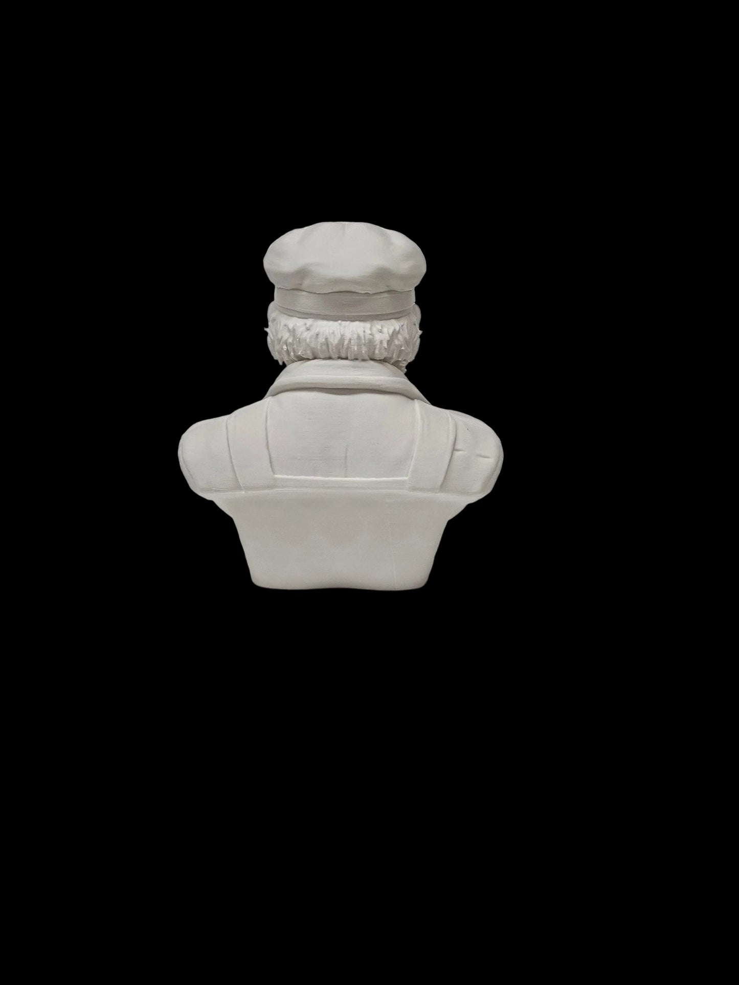 A small white ceramic figurine of a character with a cap and textured clothing, shown from the back on a white textured surface against a white speckled background.