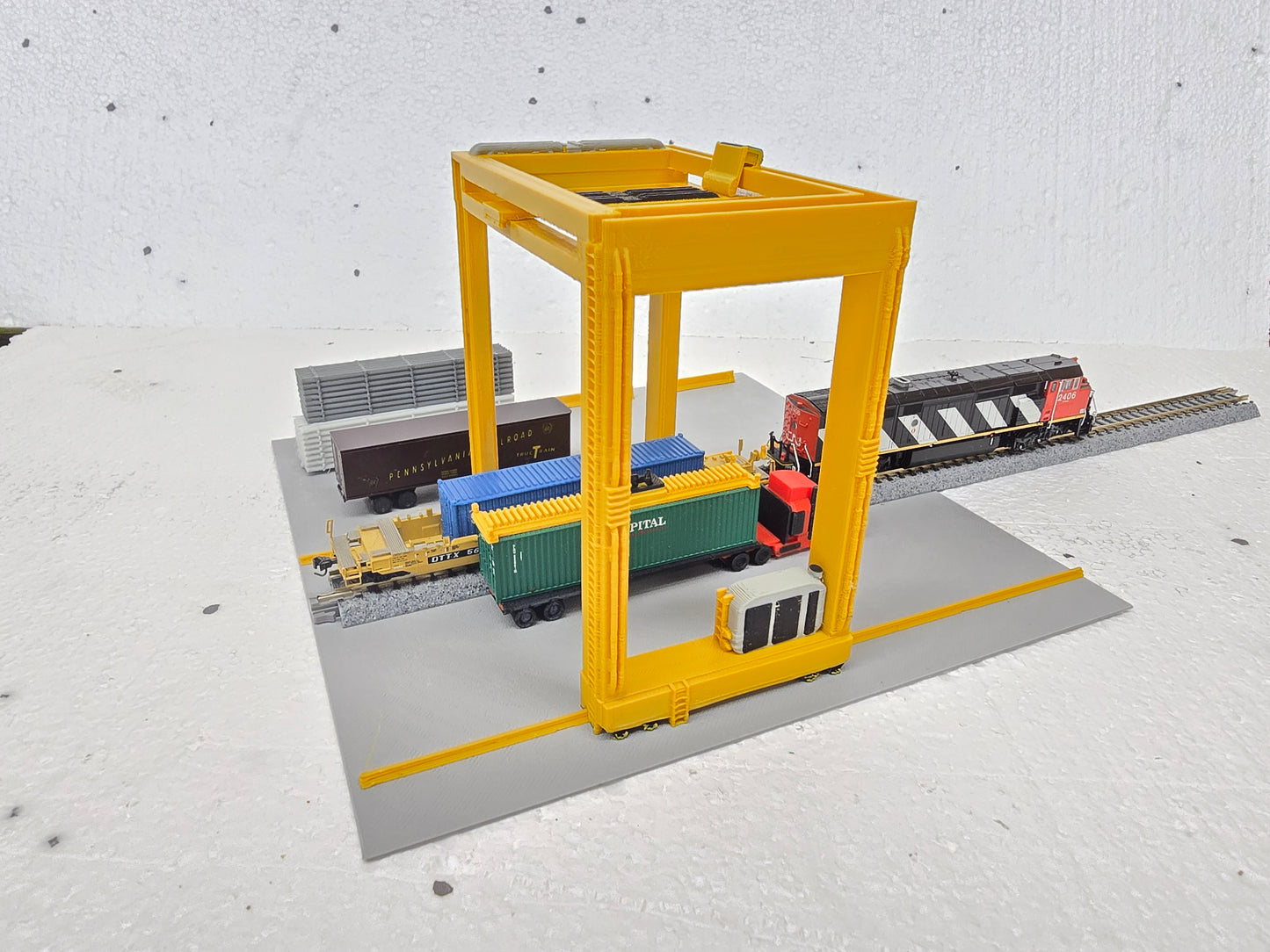 Model of a train yard with a yellow crane and toy trains on a gray surface.