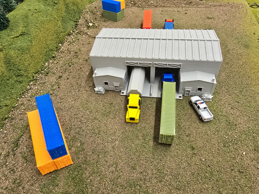 Model train layout with a gray building, yellow truck, and other miniature vehicles on a grassy surface.