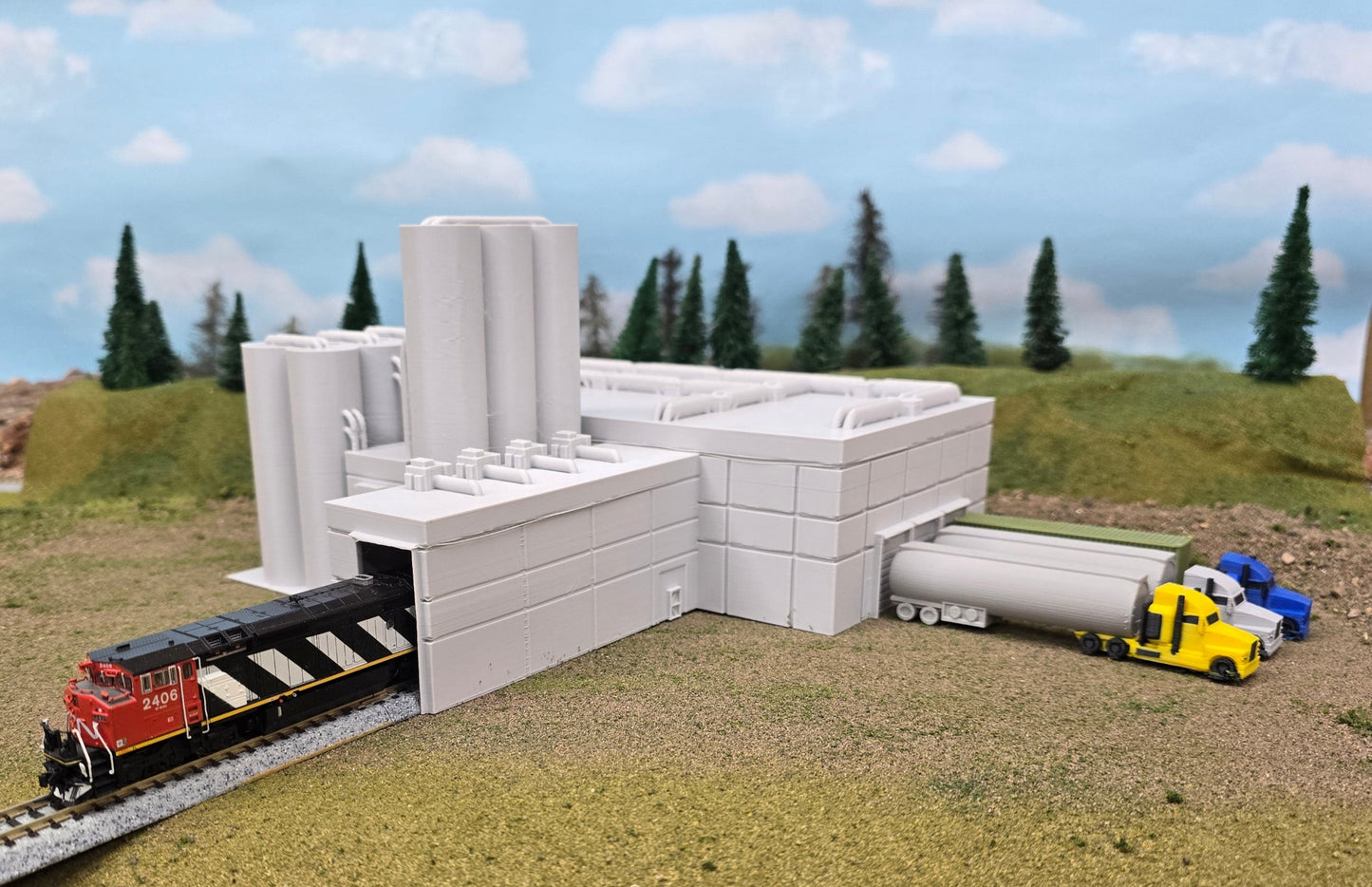 Model train set with a building, tracks, and vehicles on a grassy area with trees in the background.