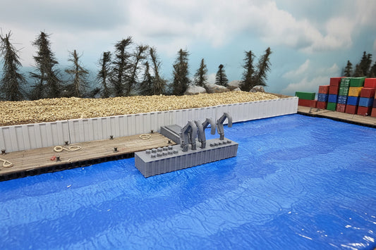 A gray 3D-printed model accessory, likely a miniature bridge or dock structure, displayed on a blue textured surface with a white fence and a miniature forest backdrop.