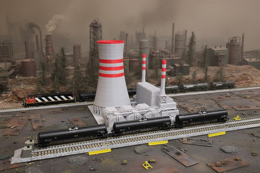 A model train set featuring a power plant with detailed structures, including a cooling tower, tanks, and a catwalk, placed on a track with surrounding landscape elements.