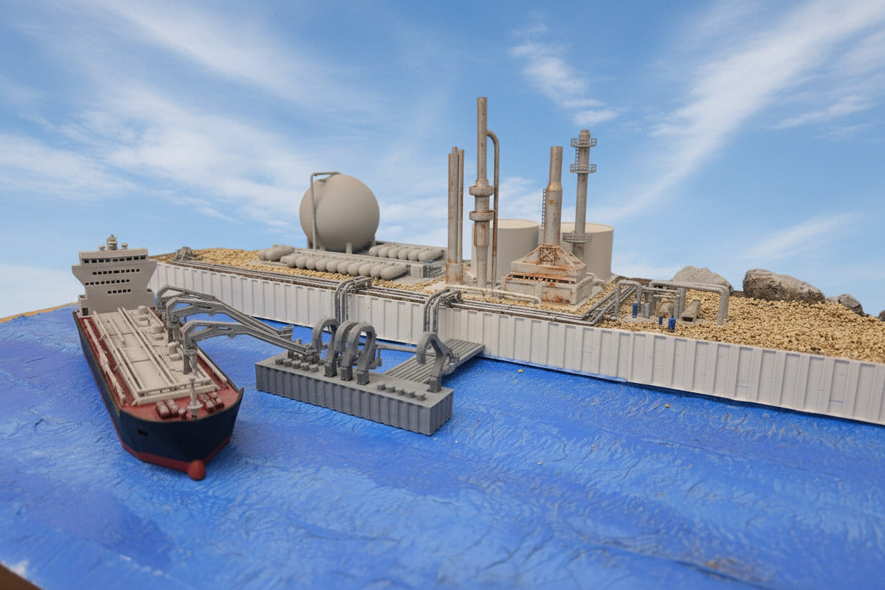 A miniature model diorama of a harbor scene, featuring a gray cargo ship and industrial dockyard structures on blue textured ground, with a painted backdrop of water, clouds, and trees.