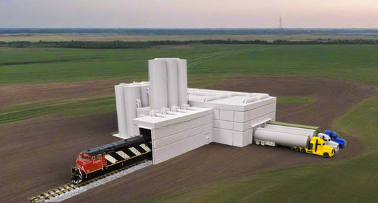 Model train set with a building, tracks, and trucks on a grassy area with trees in the background.