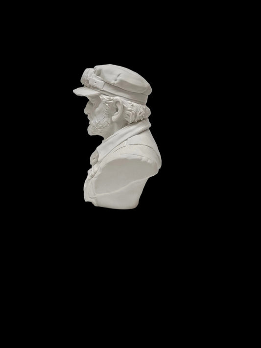 A small white bust sculpture of a bearded man in profile wearing a cap, photographed against a light textured wall and surface.