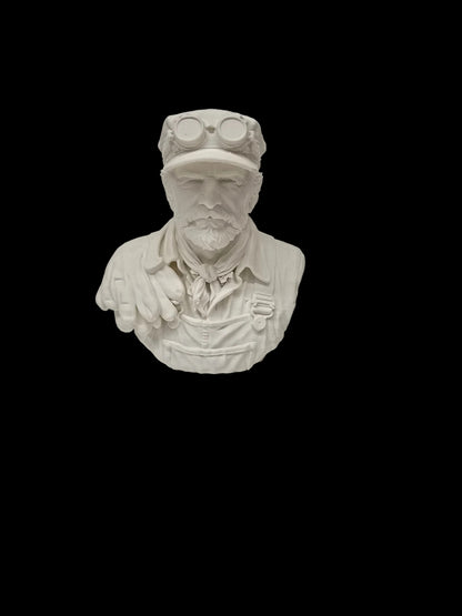 A white unpainted resin or ceramic miniature bust of a man wearing goggles on his head and a work shirt, set against a textured white background.