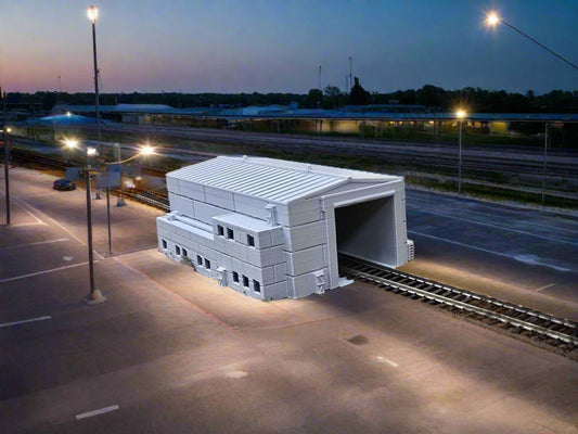 A model train set featuring a grey engine shed with detailed architecture, set against a simulated outdoor backdrop.