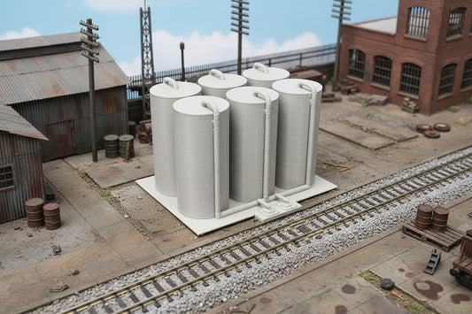 A model train storage terminal with multiple cylindrical tanks and detailed piping, set on a track with a backdrop of a desert landscape.