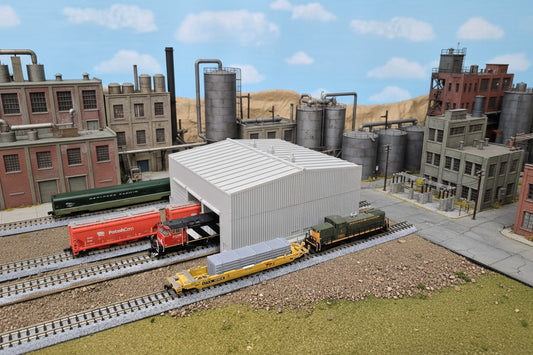 A model train set featuring a grey 5 bay train parking shed with detailed architecture, surrounded by a landscape with trees in the background.